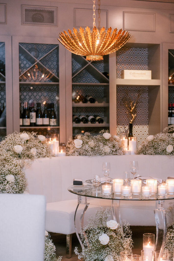 Cozy lounge seating area with a curved white banquette, glass table, and baby's breath garland beneath a gold pendant chandelier, styled by Cindy and Rita at All In Event Planning & Design.