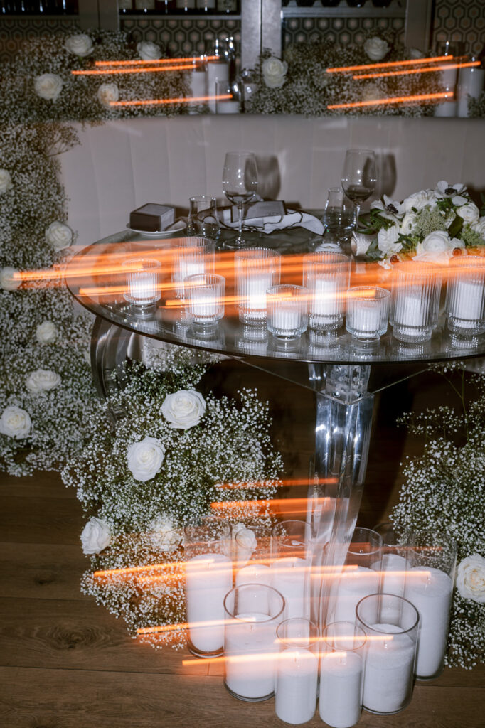 Long-exposure shot of a glass table encircled by flickering candles and baby's breath garlands, creating streaks of golden light.