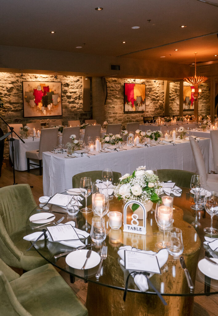 Elegant reception room with candlelit round tables, sage green velvet chairs, white floral centerpieces, and modern art on stone walls.