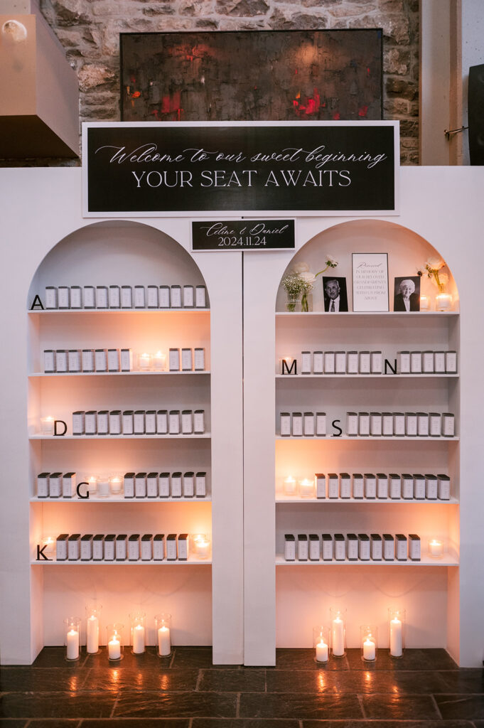 Alphabetized escort card display built into an arched wall niche, lit by candles below a "Welcome to our sweet beginning" sign, styled by Cindy and Rita at All In Event Planning & Design.