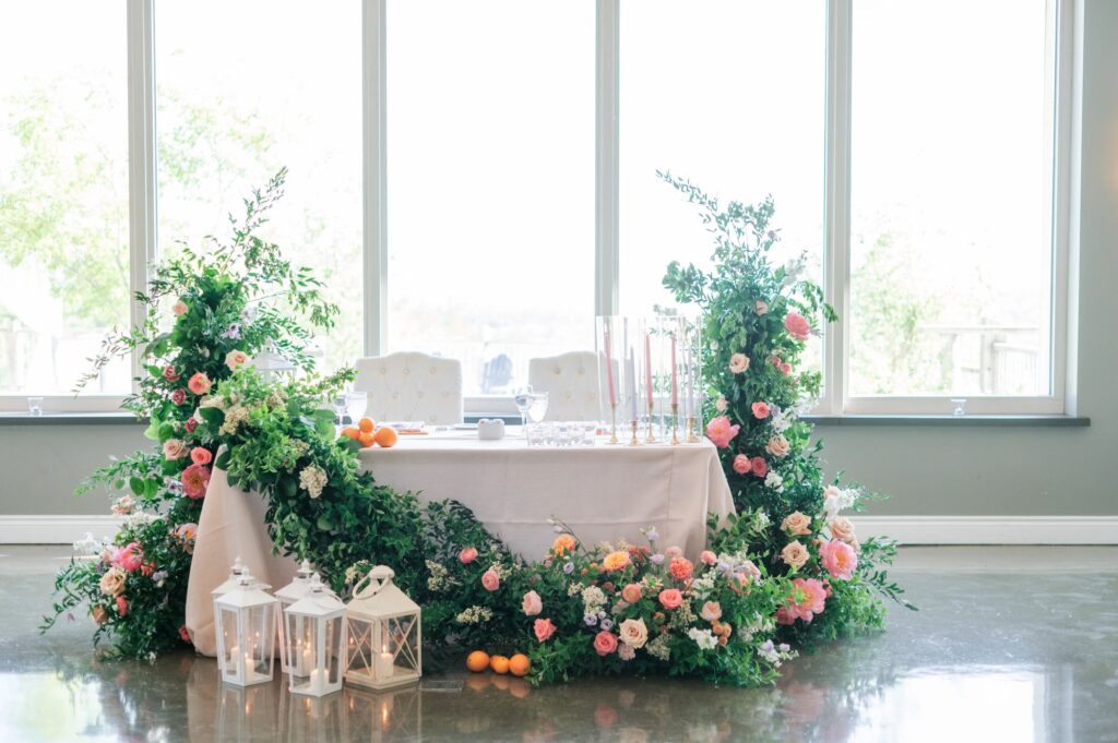 Head table draped in a garland of greenery and pink, peach, and white blooms, with white lanterns at its base and floor-to-ceiling windows behinds, planned by Amanda from Sea and Silk Events