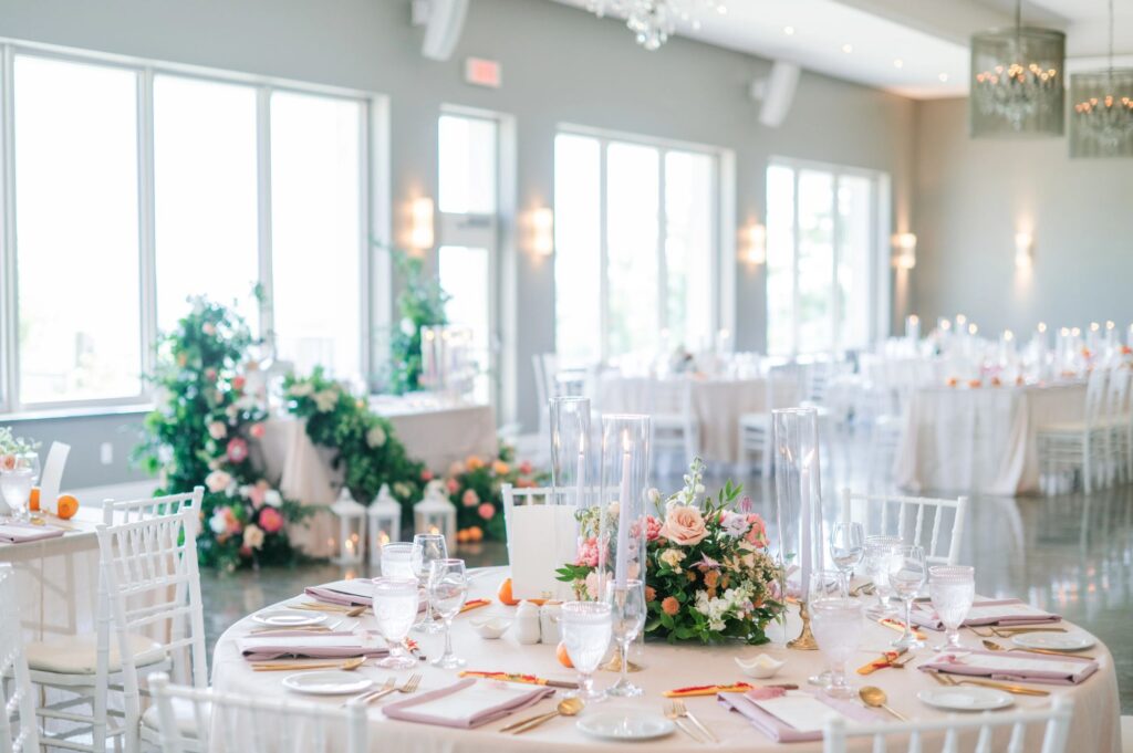 Bright wedding reception table with a low floral centerpiece in peach and pink tones, tall glass candle holders, and lantern accents, planned by Amanda from Sea and Silk Events