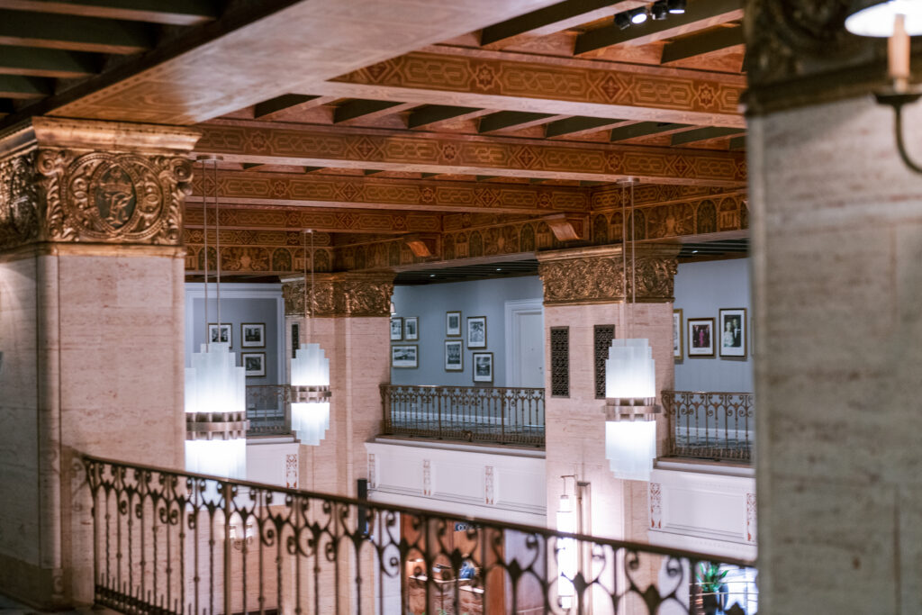 Ornate interior view of an upper mezzanine level, featuring a decorative painted wood-beam ceiling, hanging Art Deco light fixtures, wrought-iron railings, and framed artwork lining the walls below.