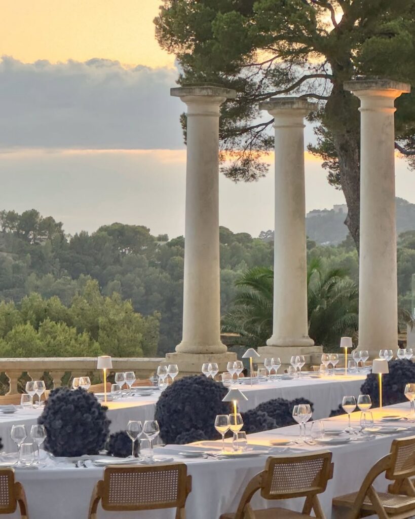Elegant outdoor dining setup with white tablecloths, blue floral centerpieces, and modern lamps. Columns and lush greenery in the background enhance a serene sunset ambiance planned by All Things Beautiful.
