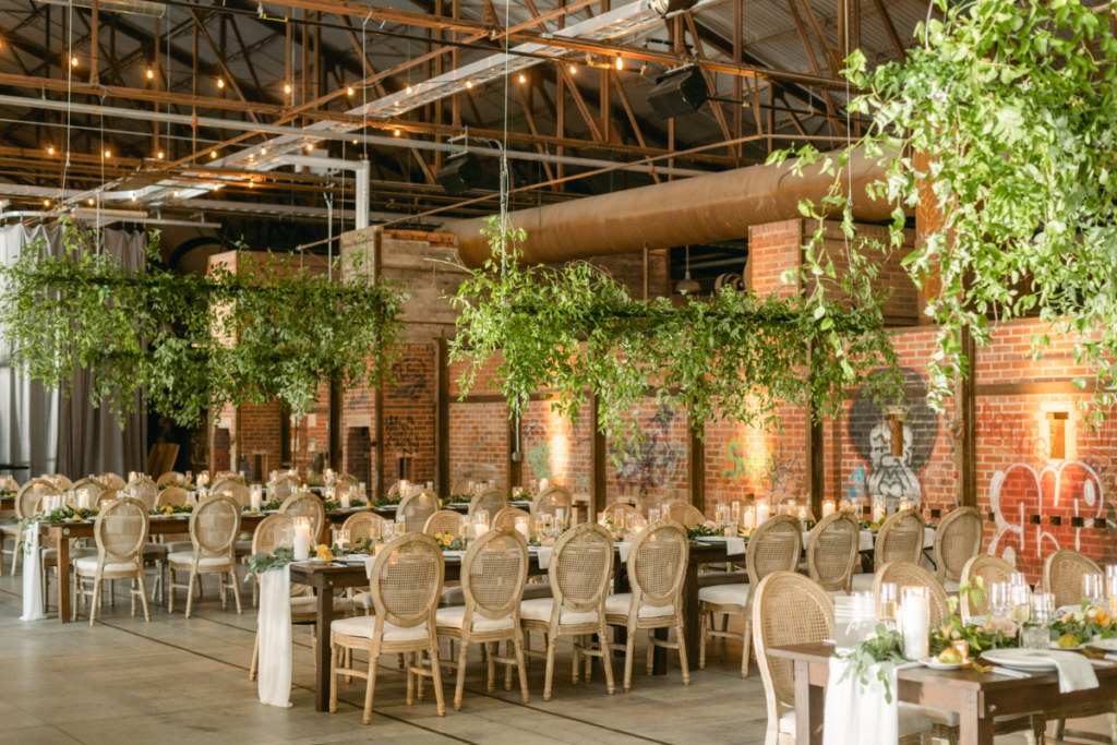 Industrial-chic wedding reception at Evergreen Brick Works in Don Valley, with cane-back chairs, greenery garlands suspended from exposed steel trusses, string lights, and exposed brick walls with graffiti accents.
