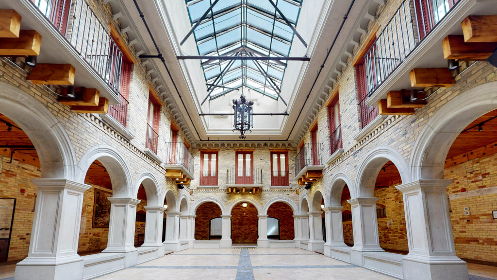 Interior courtyard at Hacienda Sereda featuring soaring white archways, brick walls with red-shuttered windows, ornate iron balconies, and a glass skylight roof.
