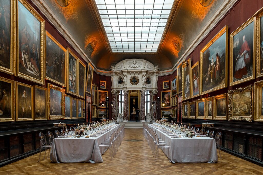 Grand gallery wedding reception set with two long banquet tables beneath ornate gilt-framed paintings and a glass skylight ceiling. Planning by Katerina Meyvial.