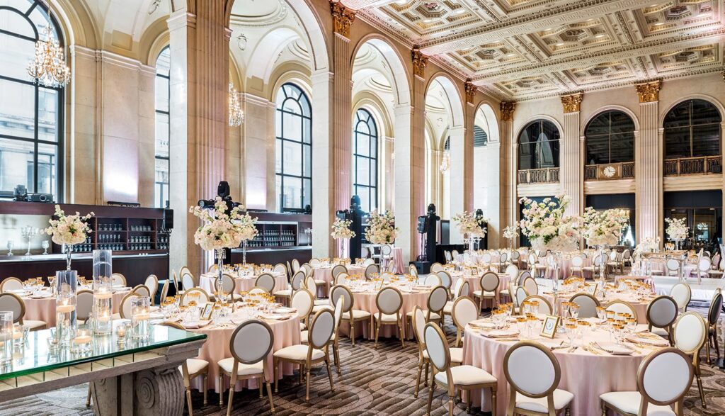 Grand wedding reception ballroom at 1 Hotel Toronto featuring round tables with blush linens, tall white floral centerpieces, and soaring arched windows beneath an ornate coffered ceiling.