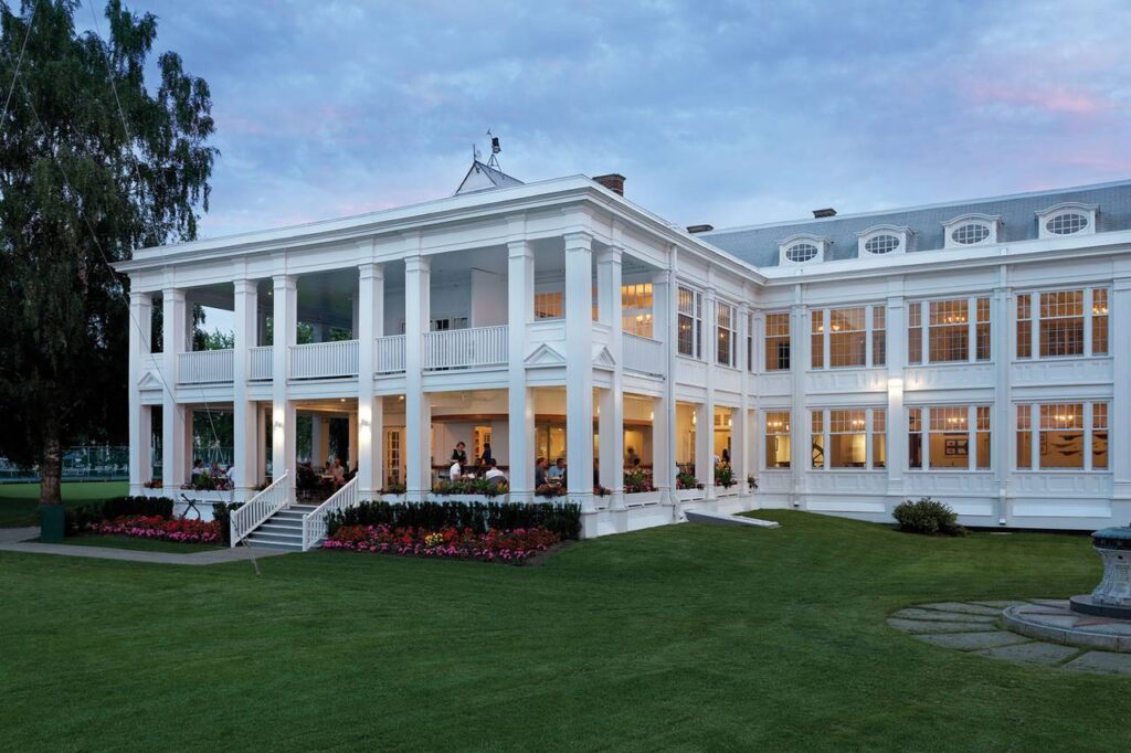 Twilight exterior view of the Royal Canadian Yacht Club on Toronto Island, a grand white colonial-style clubhouse with wraparound verandas, illuminated windows, and manicured lawns under a pastel evening sky.