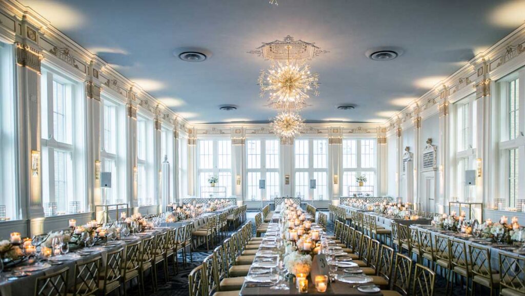 Grand ballroom wedding reception at Omni King Edward Hotel in Old Town, with long candlelit tables, gold chiavari-style chairs, ornate starburst chandeliers, and tall arched windows lining the room.