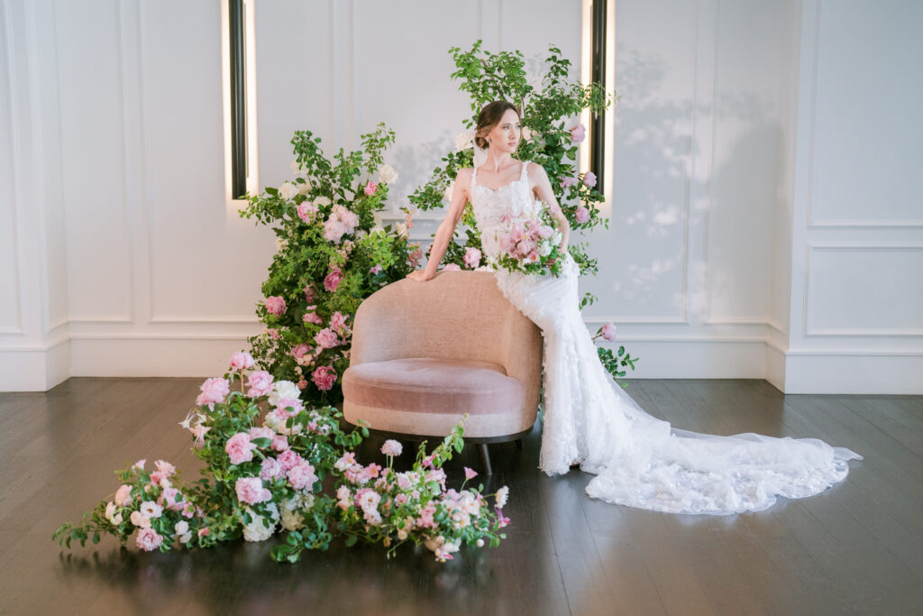 Bride in a lace wedding gown with a long train, standing beside a pink velvet chair styled with cascading pink and white floral arrangements against a white paneled wall.