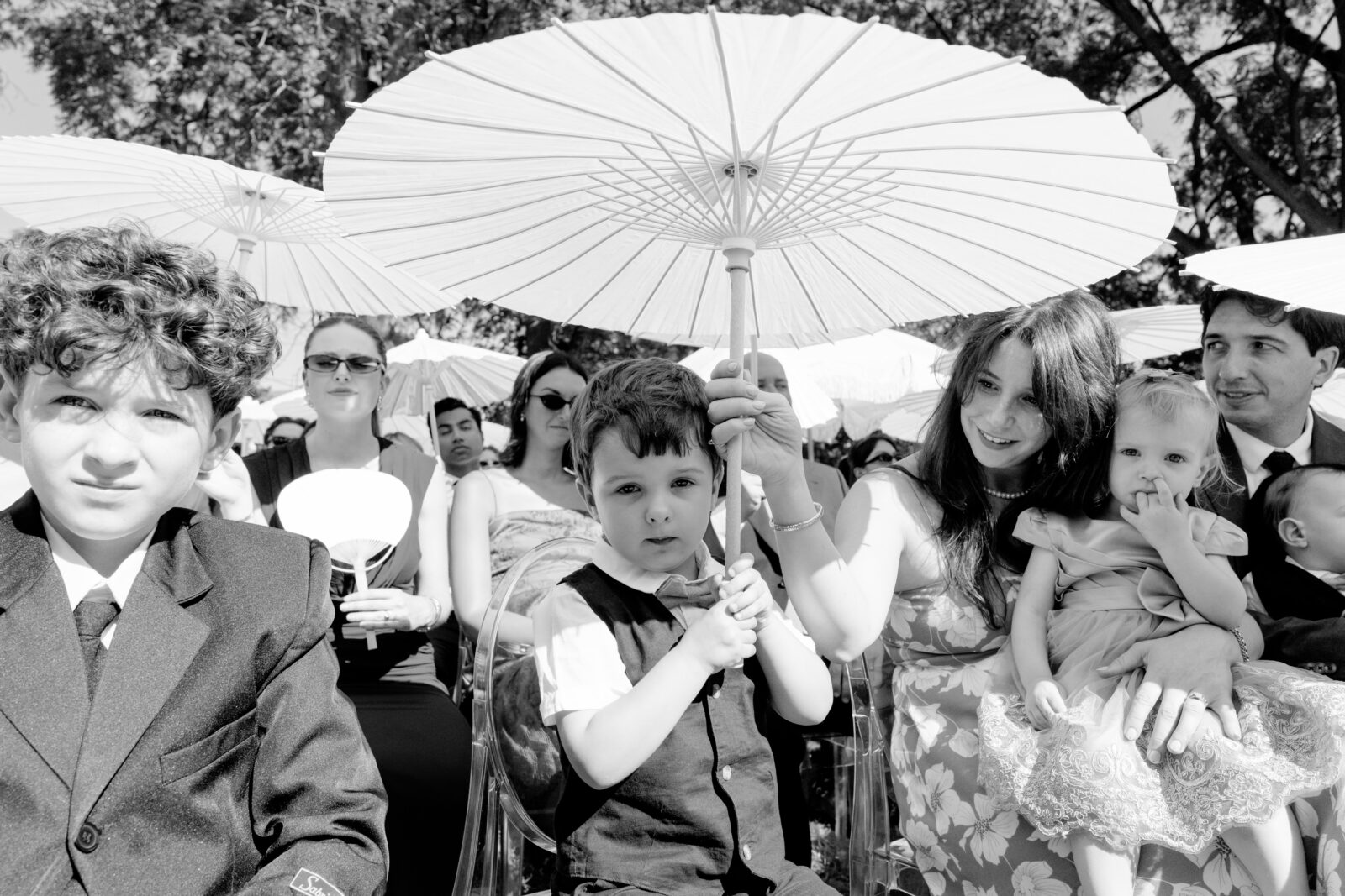 Black-and-white photo of wedding guests seated at an outdoor ceremony, with a young boy holding a white parasol in the foreground and a woman cradling a baby beside him.