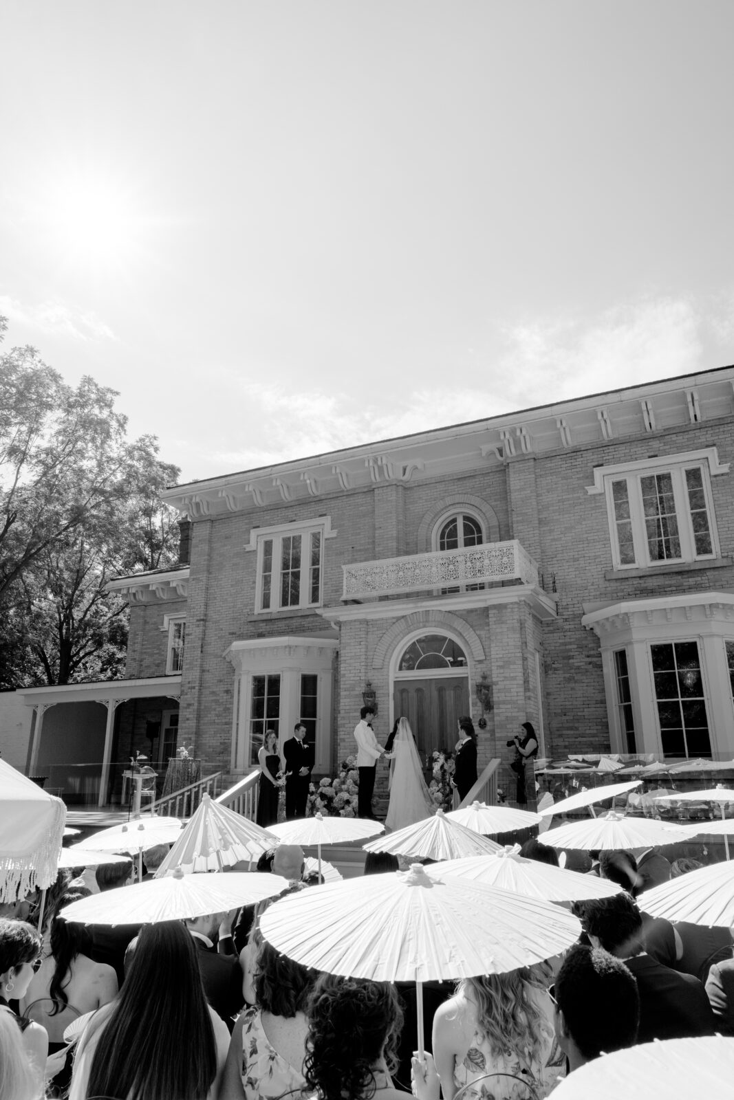 Black-and-white wide shot of an outdoor wedding ceremony at The Cape venue, with guests holding white parasols in the foreground and the couple exchanging vows on the steps of the brick manor.