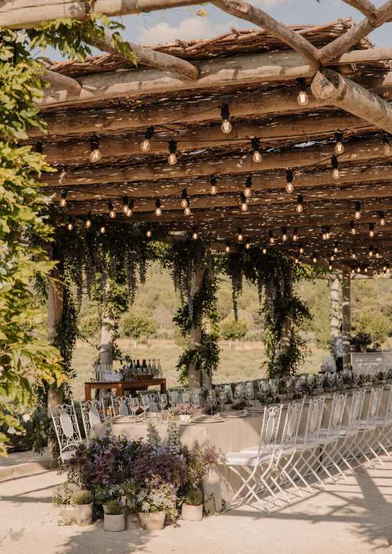 Outdoor wedding reception under a rustic wooden pergola with string lights, hanging greenery, and long tables with white chairs, planned by Ashley Pigott Events.