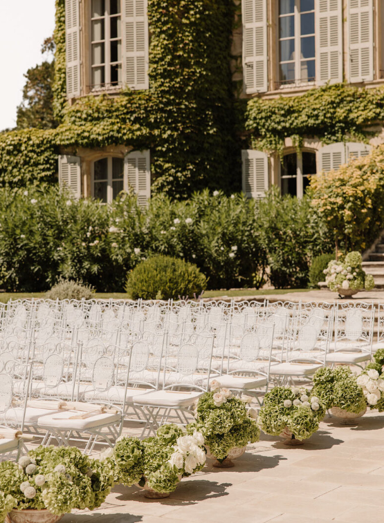 Outdoor wedding ceremony with white garden chairs and green hydrangea arrangements lining the aisle in front of an ivy-covered estate, planned by Ashley Pigott Events.