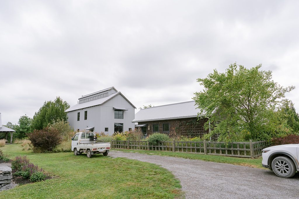 Modern grey barn and shingled cottage beside a gravel driveway and gardens at Compass Rose, a Prince Edward County wedding venue