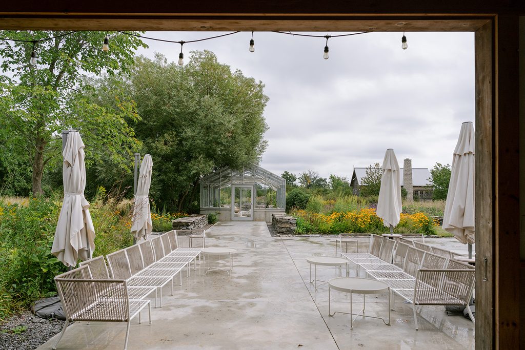 Stone patio with woven lounge chairs and string lights facing a greenhouse at Compass Rose, a Prince Edward County wedding venue