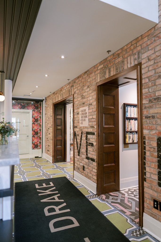 Interior entryway of Drake Devonshire featuring exposed brick walls, a patterned tile floor, and a black welcome mat with the Drake logo.