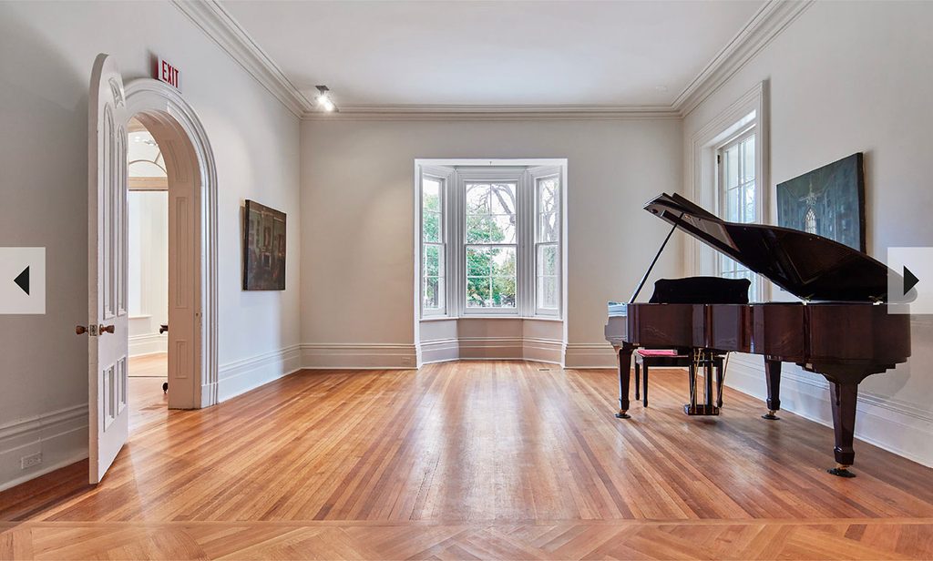Janis Joplin Hall at The Cape, a bright and airy room with herringbone hardwood floors, crown molding, arched doorways, and a grand piano by the window.