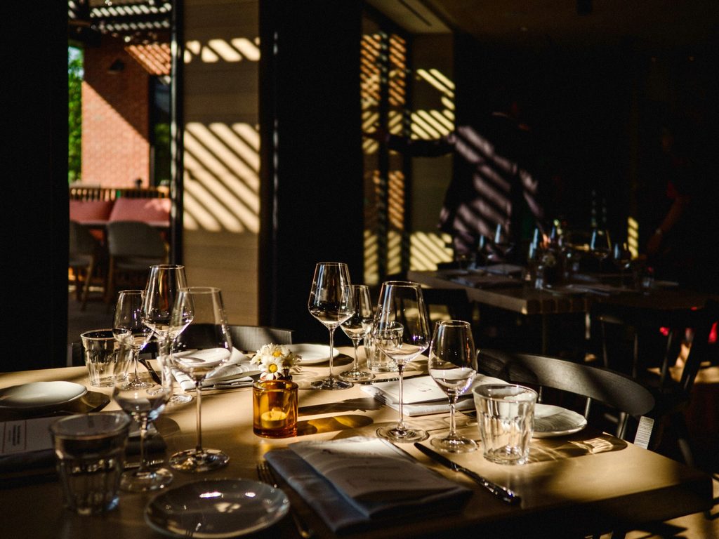 Moody, light-dappled dining table at The Royal Hotel set with crystal wine glasses, white plates, and a small amber bud vase with daisies, with dramatic lattice shadows cast across the room.