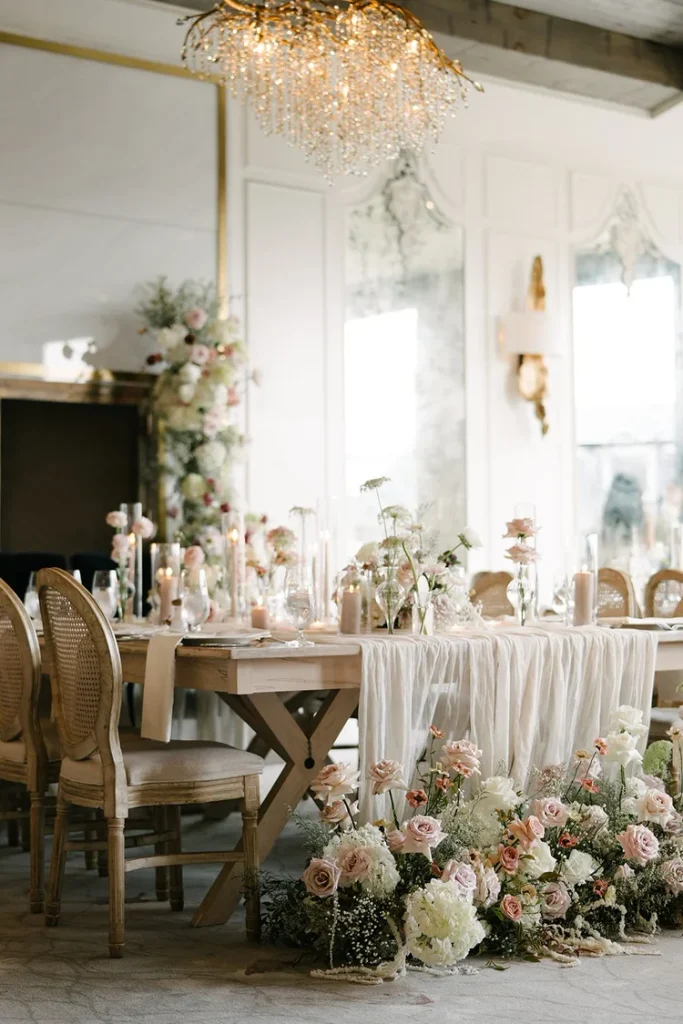 Wedding reception table with cane-back chairs, a crystal chandelier, and blush and white roses spilling onto the floor, styled by The Event Design Co, Toronto and Muskoka wedding planner.